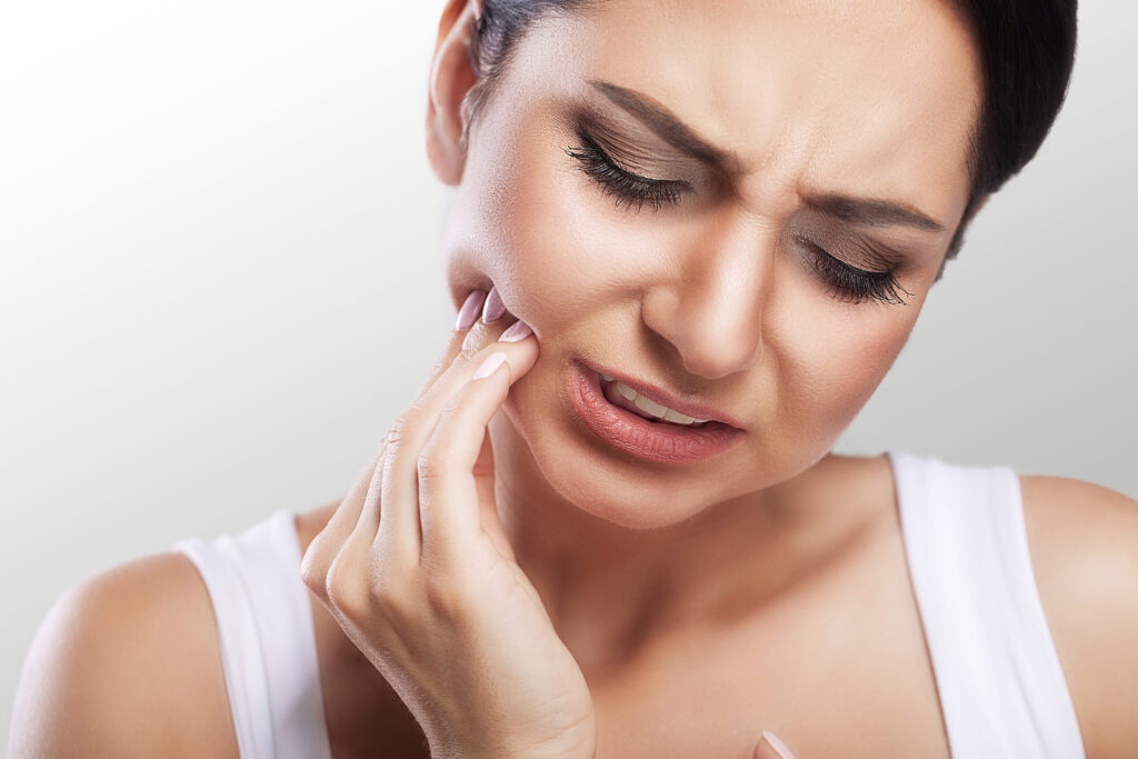 A woman holds the side of her face and grimaces due to tooth pain.