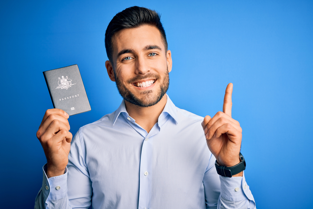 A young man holds a passport and flashes a big toothy grin while pointing upward