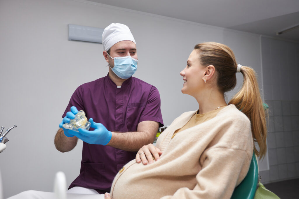 A male dentist wearing a mask and scrub cap explains treatment to a pregnant patient using a dental model.