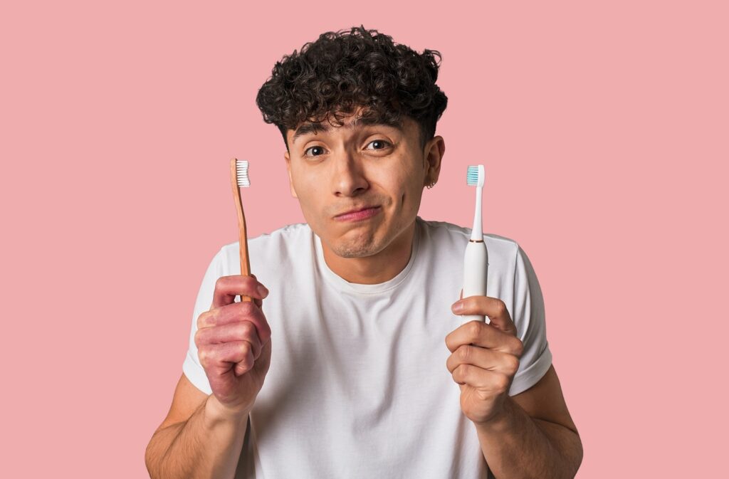 A young man holds both an electric and a manual toothbrush with a confused expression