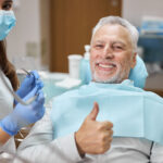 An elderly male patient giving a thumbs up to the camera while the dentist smiles beside him