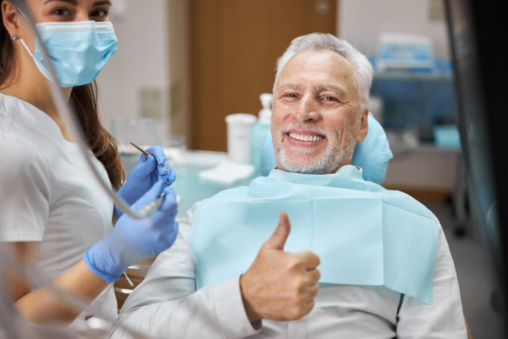 An elderly male patient giving a thumbs up to the camera while the dentist smiles beside him