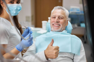 An elderly male patient giving a thumbs up to the camera while the dentist smiles beside him