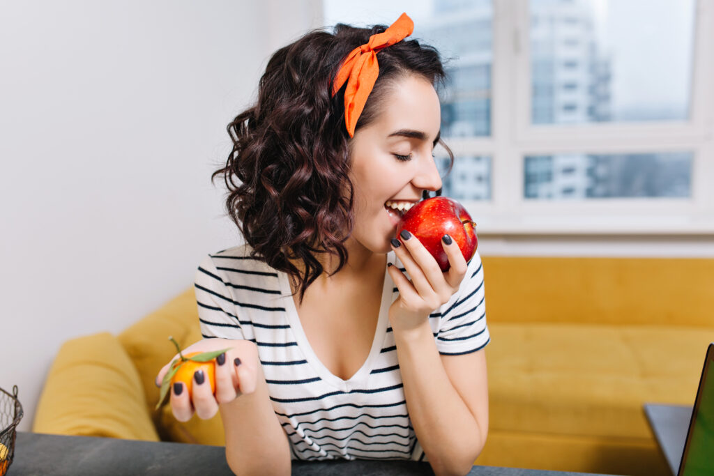 A young woman with an orange ribbon in her hair is biting into an apple and smiling joyfully.
