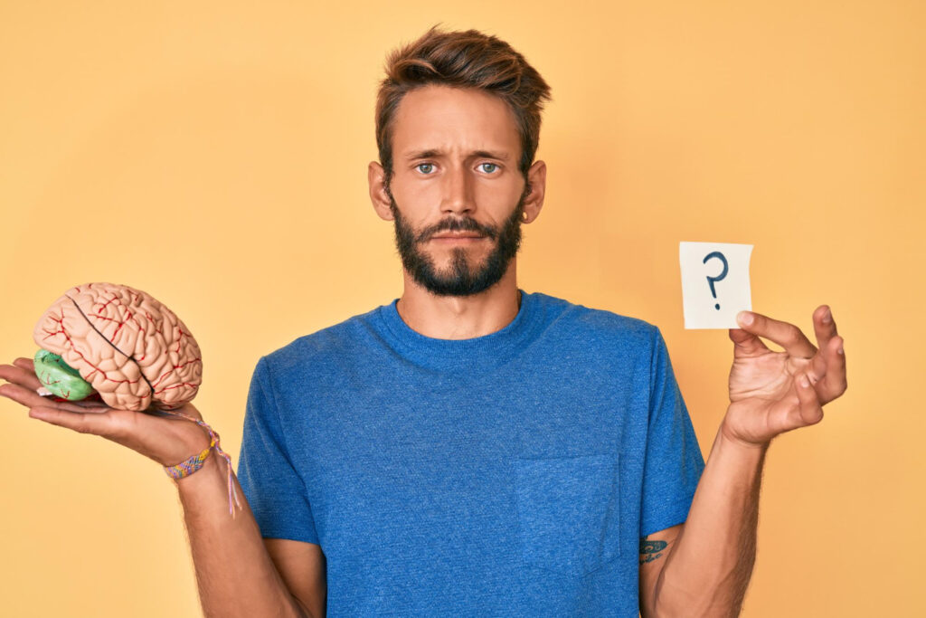 A young man with a beard holds a question mark in one hand and a brain in another