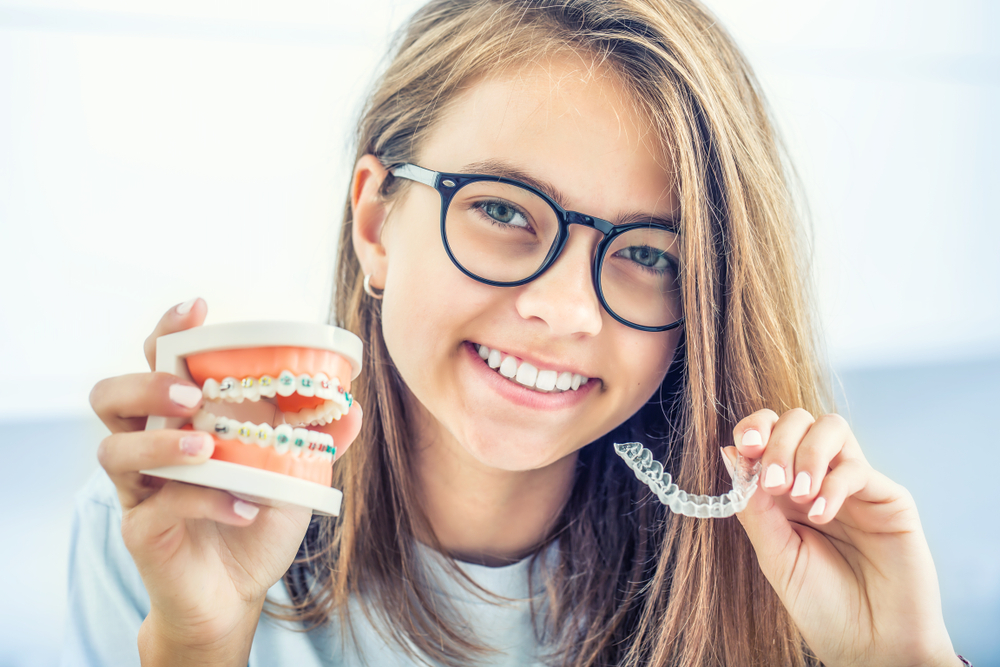A young woman holds a typodont with braces beside an Invisalign clear aligner tray