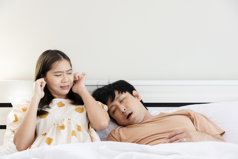 A woman covers her ears as her husband with Obstructive Sleep Apnea (OSA) snores in bed
