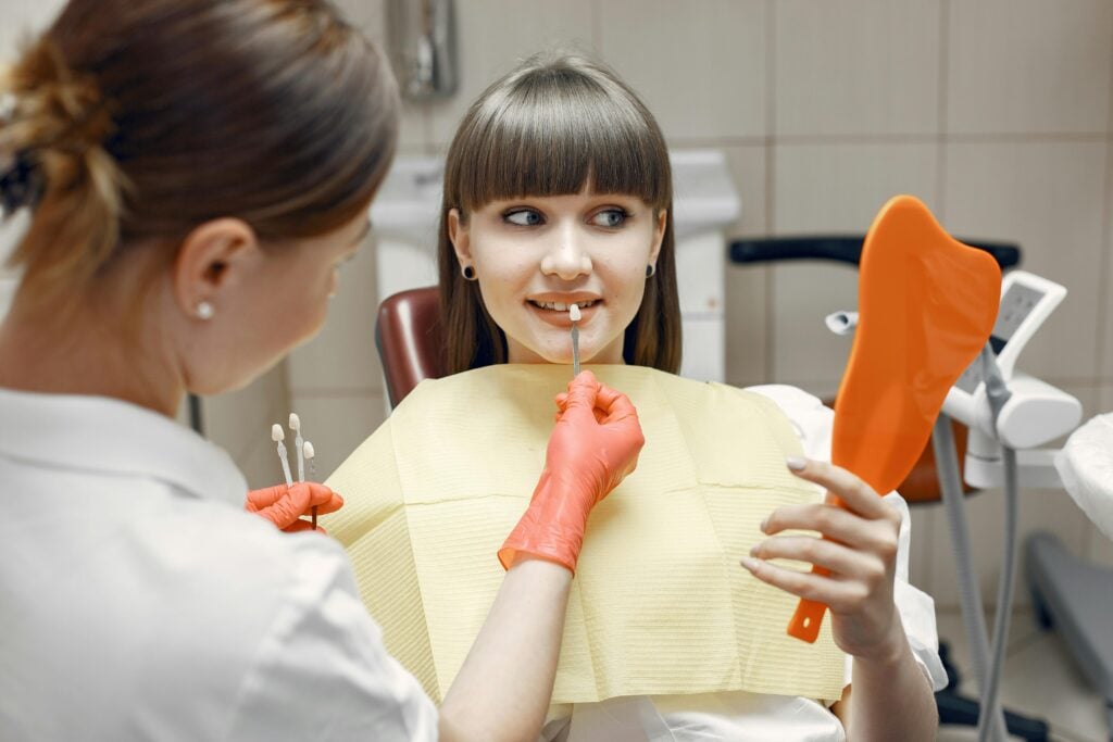 Dentist showing teeth shade samples to a patient holding a mirror.